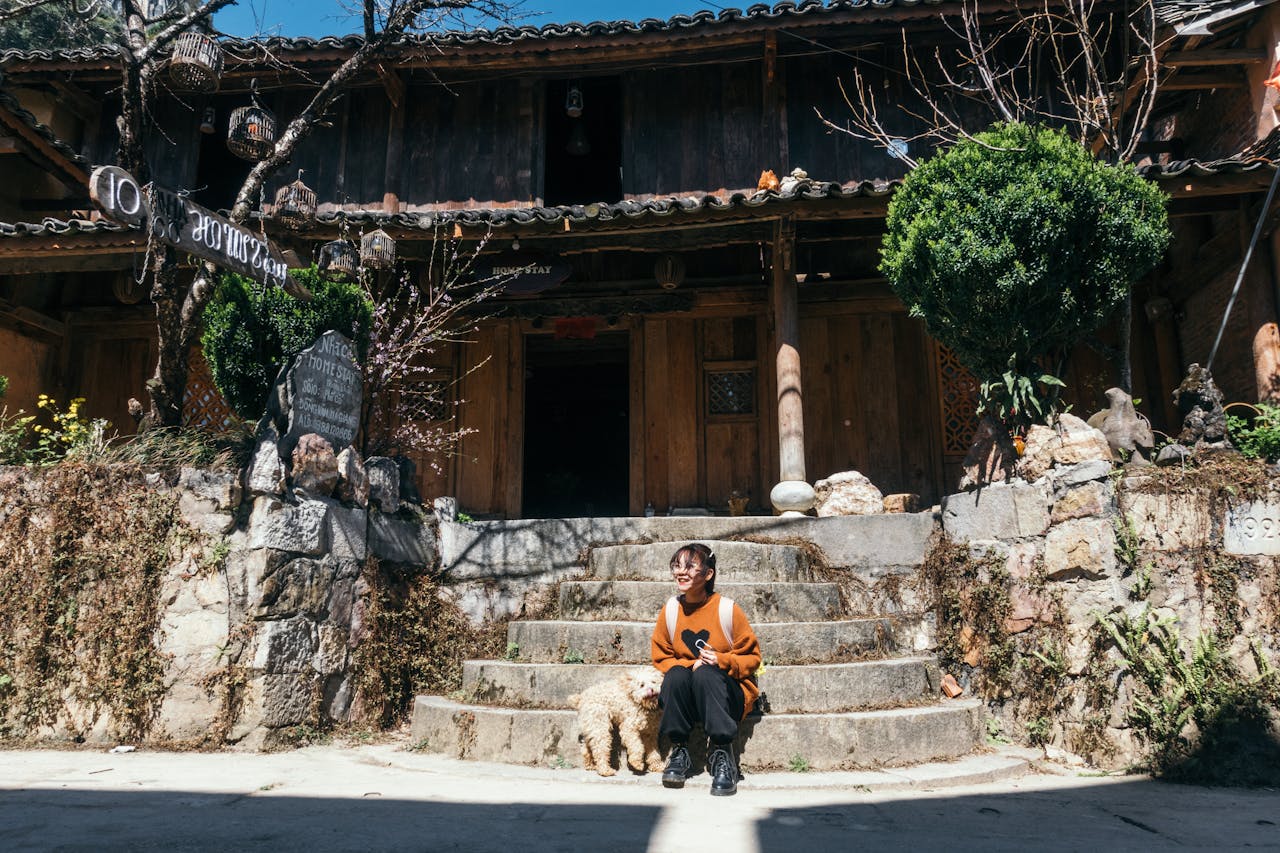 Mastering the First Impression: Your intriguing post title goes here A woman sitting on stone steps with her dog at a rustic wooden homestay entrance.